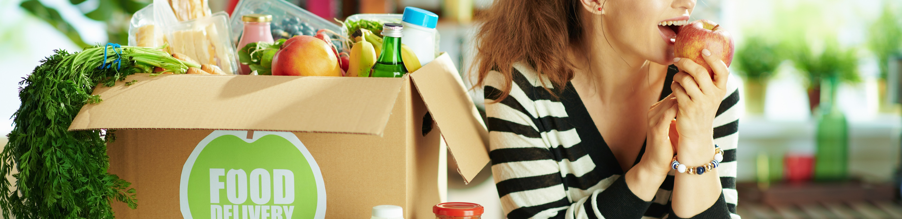Woman in kitchen receiving meal box delivery, biting into an apple